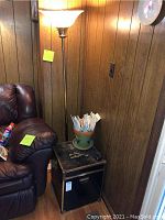 Photo showing black square trunk used as side table with brass trim and worn top, next to leather chair and floor lamp with frosted glass shade.