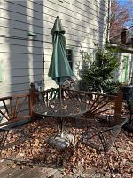 Outdoor patio scene showing round wrought iron table with green umbrella and three matching chairs surrounded by fall leaves.
