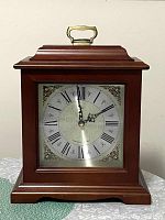 Rectangular wooden mantel clock with brass handle on top in dark brown finish, clock face has black Roman numerals on silver background with ornate floral center pattern and gold corner decoration.