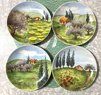 Four porcelain appetizer plates with distinct Tuscany landscape designs arranged on white fabric.