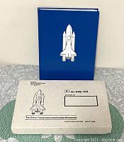Photo of the blue hardcover book with embossed space shuttle on front standing behind the original box with printed shuttle and labelling.