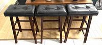 Front and top view of three dark faux leather padded bar stools with dark wood legs, showing button tufting on seats.