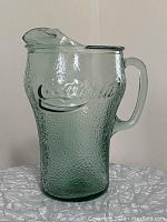 Front view of the greenish textured Coca-Cola embossed glass pitcher with handle and spout.