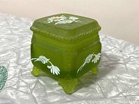 Angle view of green frosted glass trinket box showing hand painted white daisies and raised beaded edge detail. The box sits on four feet.