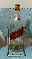 Front view of large empty Johnnie Walker Red Label bottle with attached metal cradle stand, showing label, bottle shape and cap details.