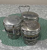 Overhead view of the four glass jelly cups with metal lids embossed with rose design, arranged in the metal carrier against a textured green mat.