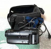 Sony Handycam Video 8 camcorder with attached strap in front of a black camera bag and power charger cable coiled in front, sitting on a green and white quilted surface.