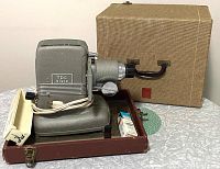 Side view showing projector with mounted lens, power cord, and brown carrying case in background along with projector lamps and 40-slide tray.