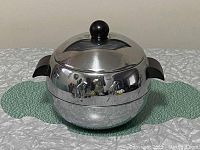 Front side view of the round chrome ice bucket with black bakelite handles and lid knob, set on a green cloth.