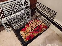 Black metal dog cage with a red and tan patterned blanket inside, gate door open, placed on tiled floor.