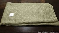 Folded green bed spread showing its color and quilt pattern on a dark wood surface with a small lot number card placed on top.