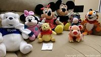 Seven Disney stuffed animals including Winnie the Pooh, Minnie Mouse, Mickey Mouse, Eeyore, and Tony the Tiger arranged on a couch.