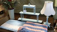 Photo showing light blue metal table lamp with pink trim, white lampshade, light blue metal shelf unit, light blue metal vase with flowers, light blue checkered bed sheet folded, and hand-knit pink and blue blanket.