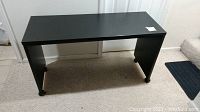 Front angled view of the black rolling table or desk showing smooth black surfaces, locking caster wheels under each leg, minor scratches visible on top surface.