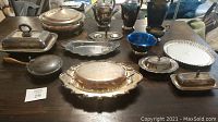 Full view of all silver-plated kitchenware items laid out on table, showing various covered serving dishes, pitchers, trays, and a blue glass bowl.