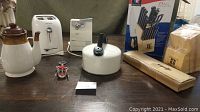 Overall shot showing small appliances, cutlery set box, kettle, coffee pot, bottle opener, and other items on table.