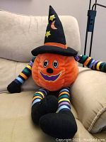 Stuffed pumpkin head plush sitting on beige couch, showing full figure with witch hat, striped limbs, and black shoes