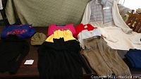 Lot of sweatshirts, sweaters, one winter jacket and hats displayed on a table and chair, varying colors: black, pink, yellow, red, grey, blue and green hat.