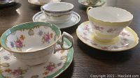Photo showing a cluster of 4 decorative teacup and saucer sets on a wooden surface including green and yellow floral patterns.