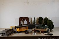 View of train station layout items showing several passenger railcars, brick tunnel portal, black metal gate with stone pillars, various trees, station platforms, and some landscape accessories.