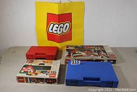 Photo of two vintage LEGO boxed sets #115 and #135 alongside red and blue storage boxes and large yellow LEGO branded bag