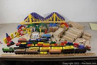 Full lot image showing wooden curved and straight track pieces stacked, yellow suspension bridge, blue truss bridge with ramps, switches, miniature road signs, trees, several wooden train engines and rolling stock lined up in front.