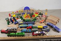 Full set display showing track assembled in oval with multiple wooden train engines and rolling stock of varied colors like red, green, blue, white, yellow, and black connected with magnets. Includes accessory pieces like buildings, trees, people, and signs surrounding the track.