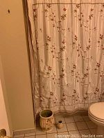Photo showing cream-colored shower curtain with floral embroidery on curtain and matching ceramic waste basket and toothbrush holder on floor.