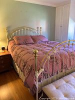 Full view of metal bed frame with headboard and footboard, showing pink bedding and part of bedroom with furniture.