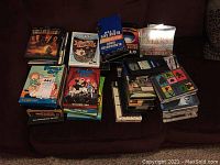 Full overview of the lot showing stacks of DVDs, VCR tapes, and CDs arranged on a couch. Includes visible Family Guy cartoon volumes, boxed DVDs, and loose VCR tapes stacked.