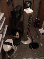 Black rotating pedestal fan, beige tower fan, small beige rotating table fan, black stand mixer with bowl in basement, surrounded by boxes and other household items.