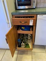 Wooden kitchen cabinet with door open displaying glass stemware including wine glasses, green decorative glass, and stainless steel travel mug.