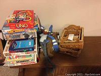Stacked board games and nested wooden baskets on a wooden surface. Games include Uno, Trouble, Game of Thrones edition, Chess and Checkers, Sorry, and Monopoly, along with a large plush toy partly visible.