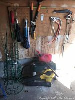 Wide view showing garden tools hanging on pegboard, green tomato cages, gas container, and yellow leaf blower on the floor.