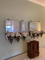 Pair of rectangular wall mirrors with glass vases and faux white roses mounted beneath. Mirrors reflect a bedroom scene.