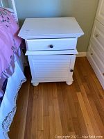 Full view of the white wooden nightstand with one drawer and one cabinet door, positioned beside a bed with pink bedding.