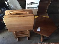 Multiple light-colored wooden TV tray tables with folding legs stored on a matching stand next to a darker wooden octagonal side table.