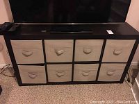 Front view of dark wood frame storage cabinet holding eight beige fabric bins with handles.