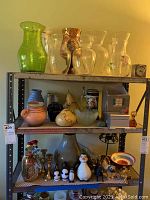 Photo of three shelves containing green and clear glass vases, ceramic jugs, jar with lid, figurines including penguins and animals, brass candlesticks and decorative bowls