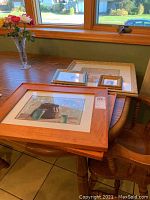 Five pieces of wall art arranged on a wooden table near window showing variety of frames and sizes