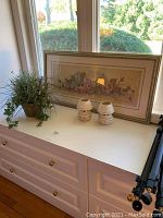 Photo of framed floral wall art, two beige candle lamps, and faux plant in container on white dresser in front of window.