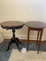 Two dark wood end tables placed side by side against a white wall and on a blue carpet. The pedestal style table has a scalloped edge top and three curved legs with brass claw feet. The other table has four simple straight legs and a round top.