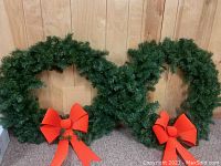 Two large green faux pine wreaths with large bright red bows set against a wooden wall.
