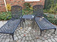 Two black wrought iron reclining lounge chairs flanking a square side table on patio pavers