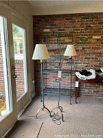 Two black wrought iron floor lamps with white lampshades standing side by side near a glass door and brick wall.