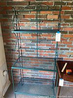 Full view of the green metal baker's rack against brick wall showing three shelves and decorative accents.