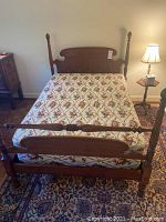 Full view of full-size bed frame with floral mattress and bedside lamp and table visible.