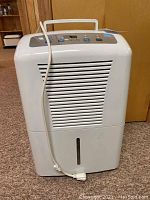 Front view of white GE dehumidifier showing body, air intake grill, and water tank window. Power cord positioned on side, plugged into unit.