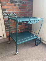 Side view of the green metal serving cart showing its two mesh shelves, floral embossed accents on the sides, and the large rear wheels with small front casters.