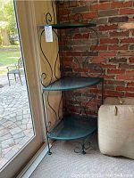Full view of the three-tier green wrought iron corner shelf in a sunroom next to a sliding glass door and brick wall.